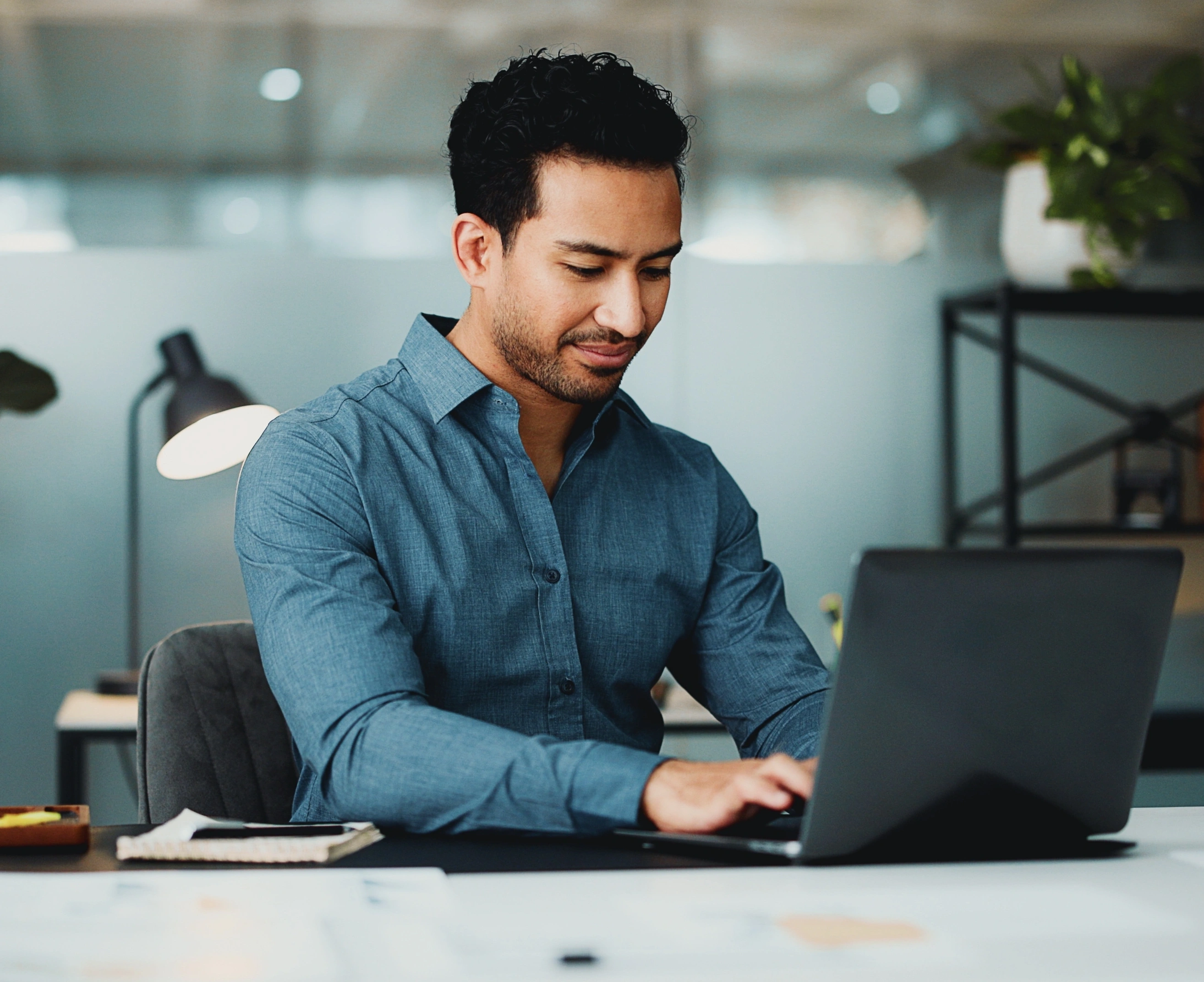 Man in a blue shirt typing on a laptop at a desk in a modern office.