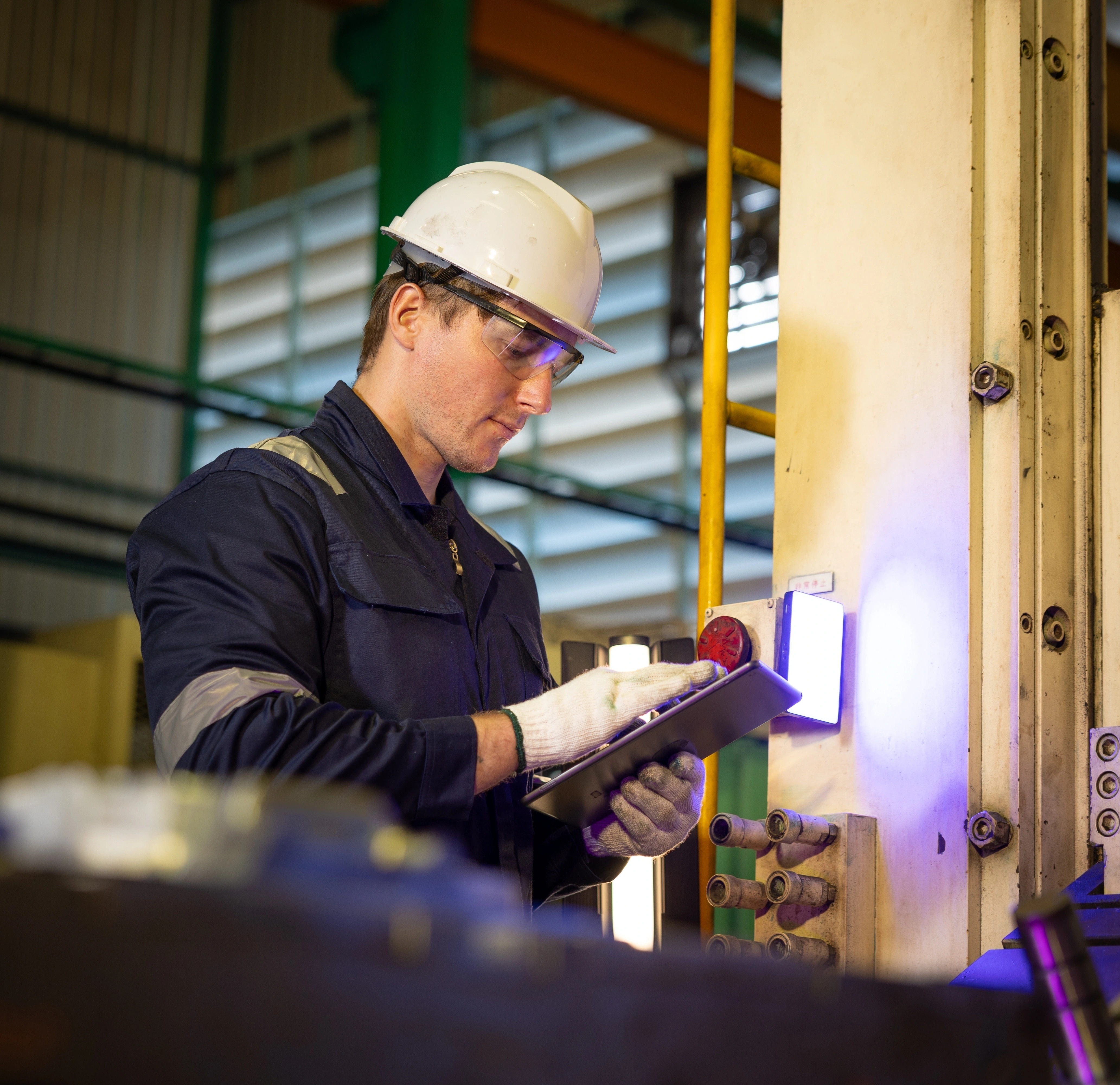 Industrial worker in a white hard hat and safety goggles using a tablet beside large industrial equipment in a factory setting