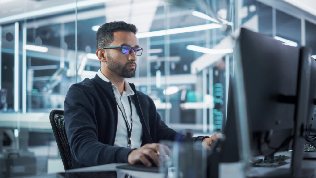 Professional man with glasses focused on a computer in a glass-walled tech office setting.