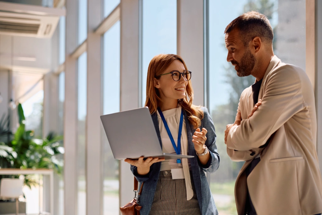 Two coworkers chat near large windows; woman holds open laptop while explaining to a man in a beige jacket.