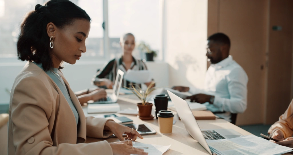 Woman in a beige blazer typing on a laptop at a light-filled office table, with two colleagues working in the background.