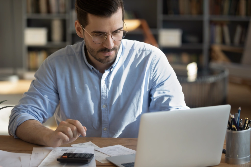 Man wearing glasses and a light blue shirt sits at a wooden desk, reviewing papers with a laptop and calculator nearby in a study or office setting.